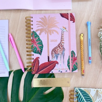 Agenda 2023 rosa com girafa e folhas tropicais, canetas e régua dourada em mesa clara