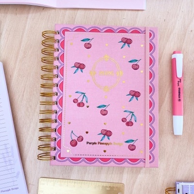 Agenda rosa com cerejas e texto dourado sobre mesa de madeira com caneta e régua