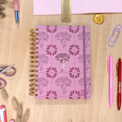 Caderno rosa floral espiral dourada com régua, canetas e clips numa mesa de madeira clara