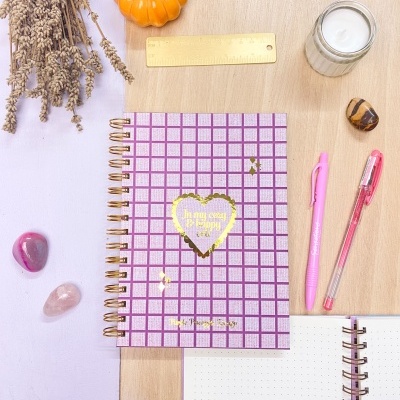 Caderno rosa quadriculado com texto dourado, régua amarela, canetas cor-de-rosa, vela em vidro, abóbora cerâmica laranja, ramos secos e pedras decorativas sobre mesa