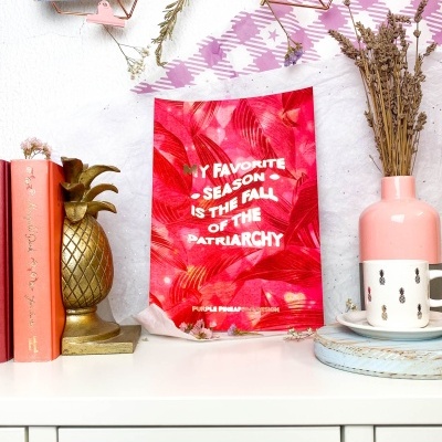 Prateleira com livros cor de rosa, escultura dourada ananás, cartaz vermelho com texto branco e vaso rosa com flores secas.