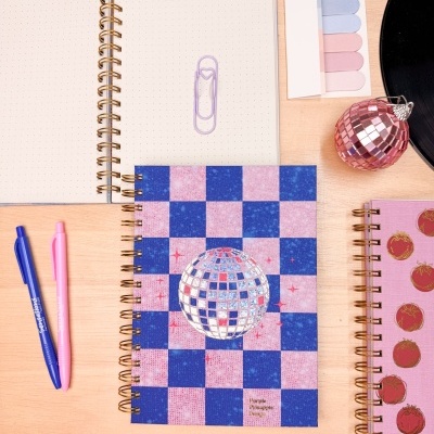 Cadernos espiral com capas decoradas em padrão xadrez e imagens de tomates, canetas, marcadores de página pastel e objeto decorativo rosa numa mesa de madeira.