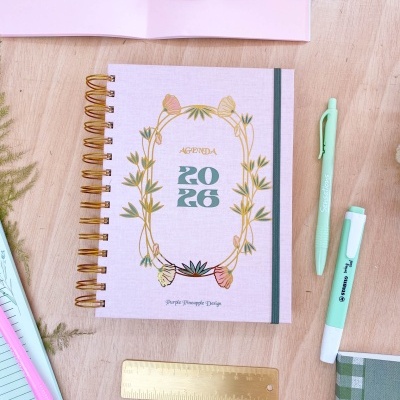 Agenda 2026 com capa rosa e decoração de plantas, caneta, marcador e régua sobre madeira