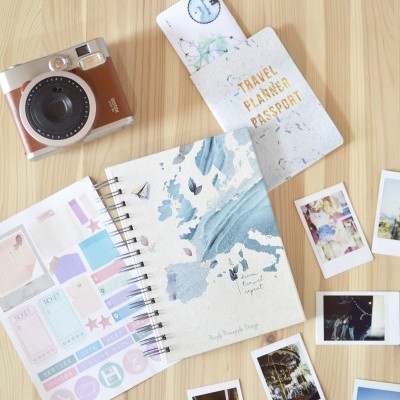 Câmara vintage, caderno de capa azul e bege, autocolantes coloridos, fotografias instantâneas e passaporte sobre madeira clara