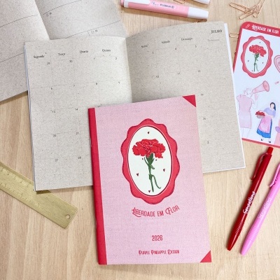 Agenda 2028 rosa com desenho de flor e texto LIBERDADE EM FLOR, aberta, com marcadores e canetas numa mesa de madeira.