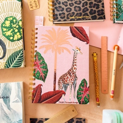 Cadernos decorativos espirais com padrões animais e tropicais ao lado de réguas e canetas numa mesa.