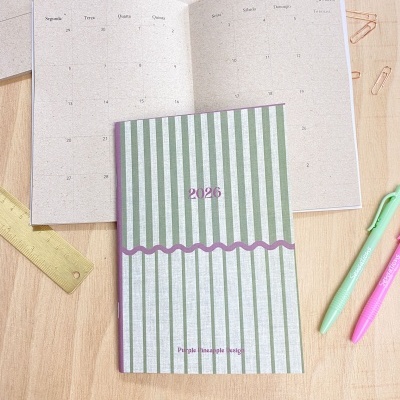 Agenda 2026 com capa verde e branca às riscas, aberta em calendário mensal com canetas e régua sobre mesa