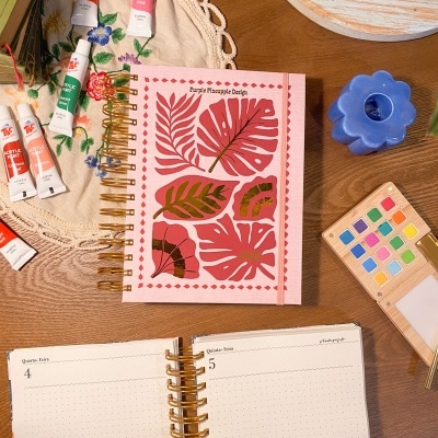 Agenda rosa com folhas, tubos de tinta acrílica, paleta de aquarela, vela azul em flor, mesa de madeira