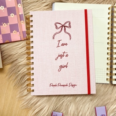 Caderno rosa claro com texto em vermelho e elástico vermelho, rodeado de outros cadernos e marcadores