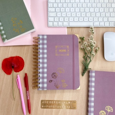 Caderno roxo 2026 com padrão xadrez e detalhes dourados na mesa com teclado, canetas e flores