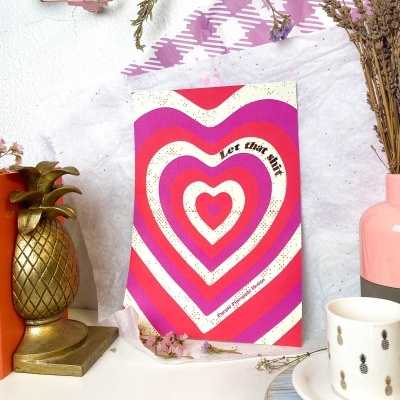 Livro com capa colorida em formato de corações, vaso rosa com flores secas, caneca branca com abacaxis, suporte dourado em forma de abacaxi numa bancada branca