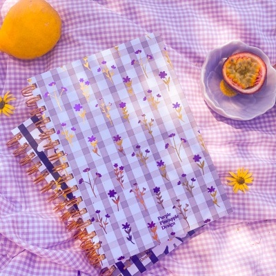 Cadernos com capas xadrez lilás e branco com flores pequenas e texto Purple Pineapple Design sobre tecido xadrez lilás com fruta passion fruit e laranja