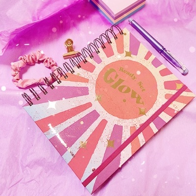 Caderno colorido com texto Ready Set Glow e acessórios de escritório em fundo rosa