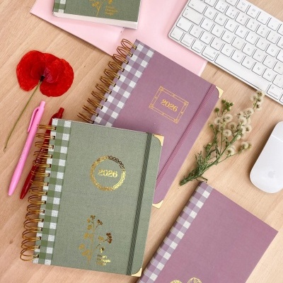 Cadernos com capas verdes e lilás com detalhes dourados e padrão xadrez, caneta rosa, flores, teclado e rato branco sobre mesa de madeira