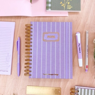 Agenda 2025 lilás às riscas com canetas e régua sobre mesa de madeira