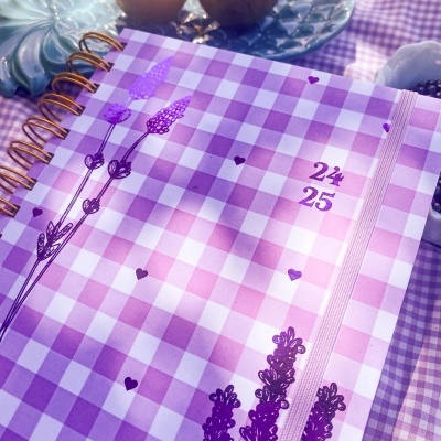 Caderno lilás xadrez com flores e números 24 e 25, ao lado de maracujás em prato azul