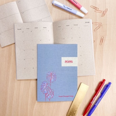 Agenda 2026 azul com ilustração vermelha, canetas, clips e marcador sobre mesa de madeira