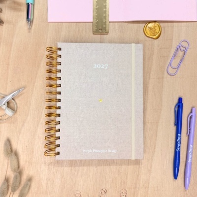 Calendário ou agenda 2027 com capa bege texturada e espiral dourada numa mesa com material de escritório.