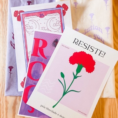 Sacos de tecido lilás e bege com flores e três cartazes coloridos sobre madeira clara, incluindo cartaz com a palavra RESISTE e ilustração de cravo vermelho