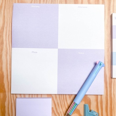 Material de escritório com bloco de notas quadrado lilás e branco, caneta colorida, clipes e amostras de cores sobre madeira clara