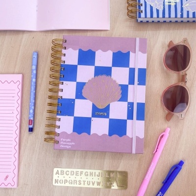 Caderno espiral azul e lilás com canetas, etiquetas, régua e óculos de sol