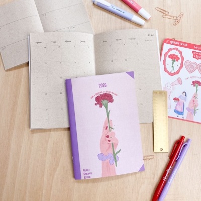 Agenda ilustrada com canetas, régua dourada, clips e autocolantes numa mesa de madeira clara