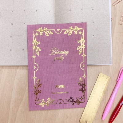 Agenda rosa 2026 decorada com dourado sobre mesa de madeira com canetas e régua