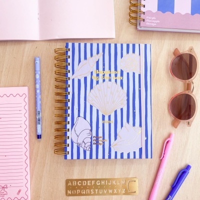 Caderno azul às riscas com conchas, canetas, régua de alfabeto e óculos de sol sobre mesa de madeira