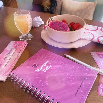 Mesa com caderno rosa, caneta, copo de bebida e tigela com frutas, cão ao fundo no sofá