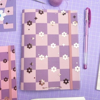 Caderno rosa e lilás com padrão xadrez e flores em relevo numa mesa lilás com acessórios de artesanato