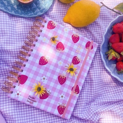 Caderno espiral roxo com morangos e flores, rodeado por limões, morangos e maracujás sobre tecido roxo aos quadrados.