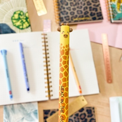 Lápis amarelo com padrão de girafa e texto 'HEAD IN THE CLOUDS' sobre mesa com cadernos e réguas