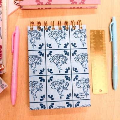 Caderno azul claro com padrão de flores e gotas em espiral dourada, régua dourada e canetas cor-de-rosa e azul-claro