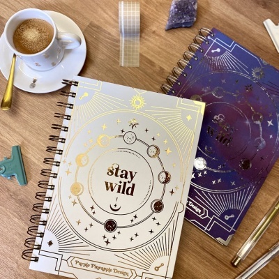 Duas agendas espirais com desenhos dourados e uma chávena de café sobre uma mesa de madeira.