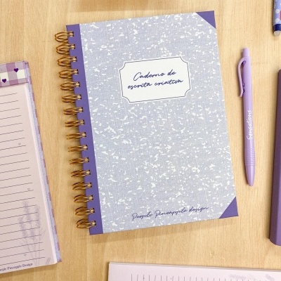 Caderno de escrita criativa com capa cinza e roxa, espiral dourada e caneta roxa sobre mesa de madeira.