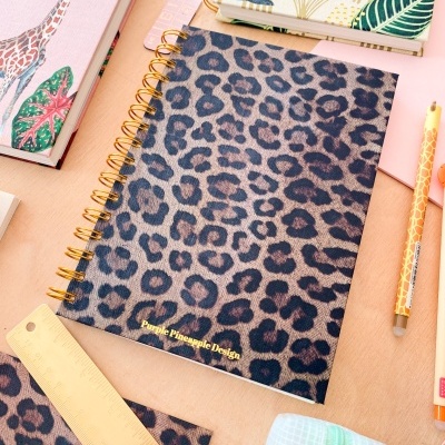 Caderno com capa animal print e espiral dourada ao lado de régua, lápis e outros cadernos coloridos