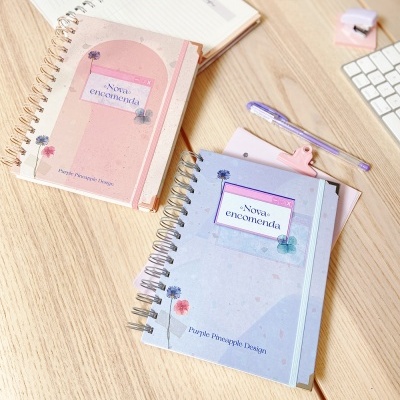 Dois cadernos espirais rosa e azul com textos e flores numa secretária de madeira com acessórios de escritório