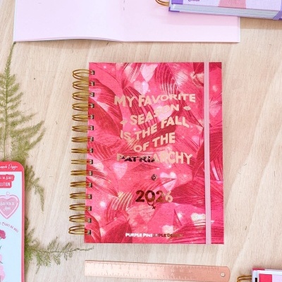 Caderno espiral vermelho com padrão floral e texto dourado sobre mesa clara