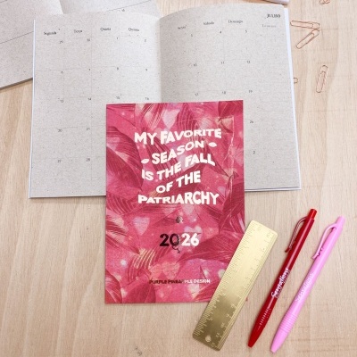 Planner 2026 com capa vermelha e padrão floral, texto branco, aberto sobre mesa de madeira com materiais de escrita