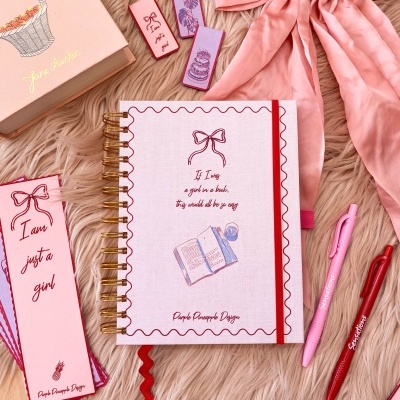 Diário rosa claro com ilustração e texto, canetas, marcadores de página, laço rosa e caixa com ilustração, sobre superfície felpuda
