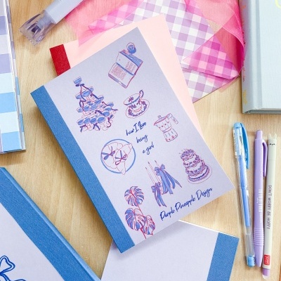 Caderno com capa azul e ilustrações coloridas, canetas pastel, fita rosa e envelope xadrez sobre mesa de madeira clara.