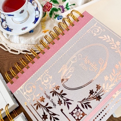 Agenda 2026 com capa cinza e rosa com detalhes florais dourados ao lado de chávena de chá em porcelana e pano bordado