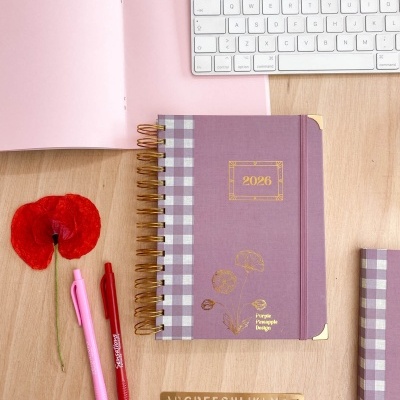 Agenda 2026 roxa com detalhes dourados, canetas, flor vermelha, teclado branco, capa rosa aberta e régua alfabética sobre mesa de madeira