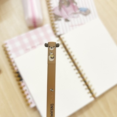 Caneta castanha com ponta em forma de ursinho e cadernos espiralados abertos sobre mesa