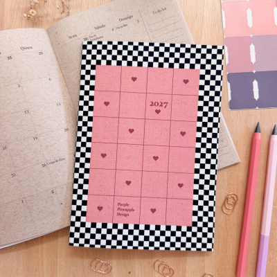 Agenda 2027 com capa xadrez preto e branco e quadrado rosa com corações, sobre mesa de madeira com caderno aberto, clips e lápis.