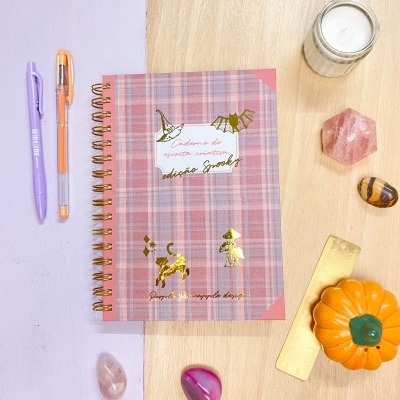 Caderno quadriculado rosa com detalhes dourados e canetas, vela, pedras e régua numa mesa