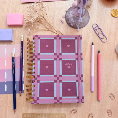 Caderno 2027 rosa e roxo com padrão xadrez e corações, lápis, clips, envelopes selados, régua de madeira e vela derretida.