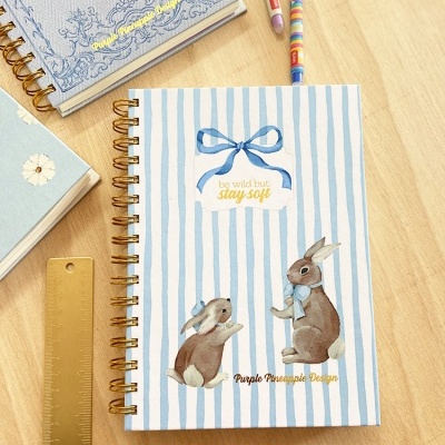 Cadernos com espiral dourada, um com coelhos e riscas azuis, régua dourada e lápis sobre mesa clara