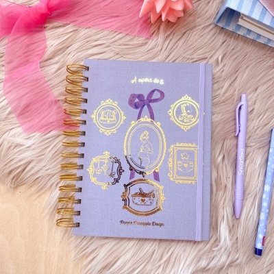 Caderno lilás decorado com molduras douradas, fita rosa e objetos ao redor