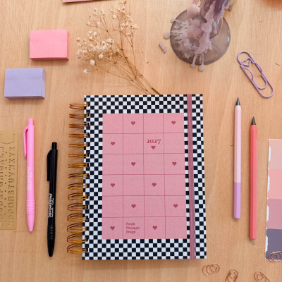 Caderno preto e branco com rosa, canetas, clips e vela rosa derretida num fundo de madeira.